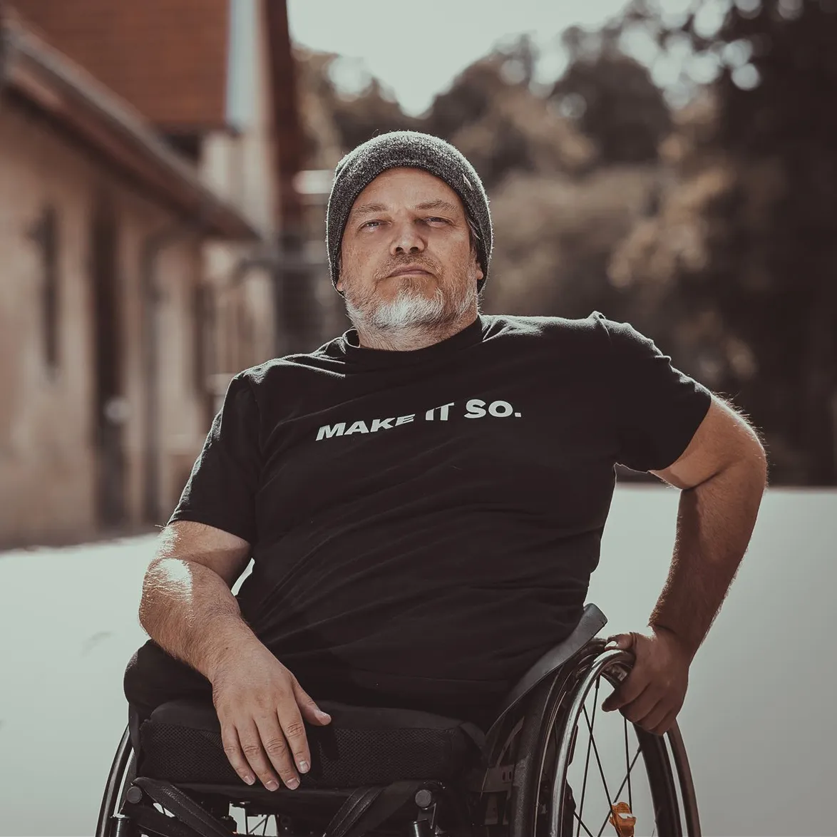 Macs Moser outdoors in his wheelchair, in a beanie and a black t-shirt reading MAKE IT SO.